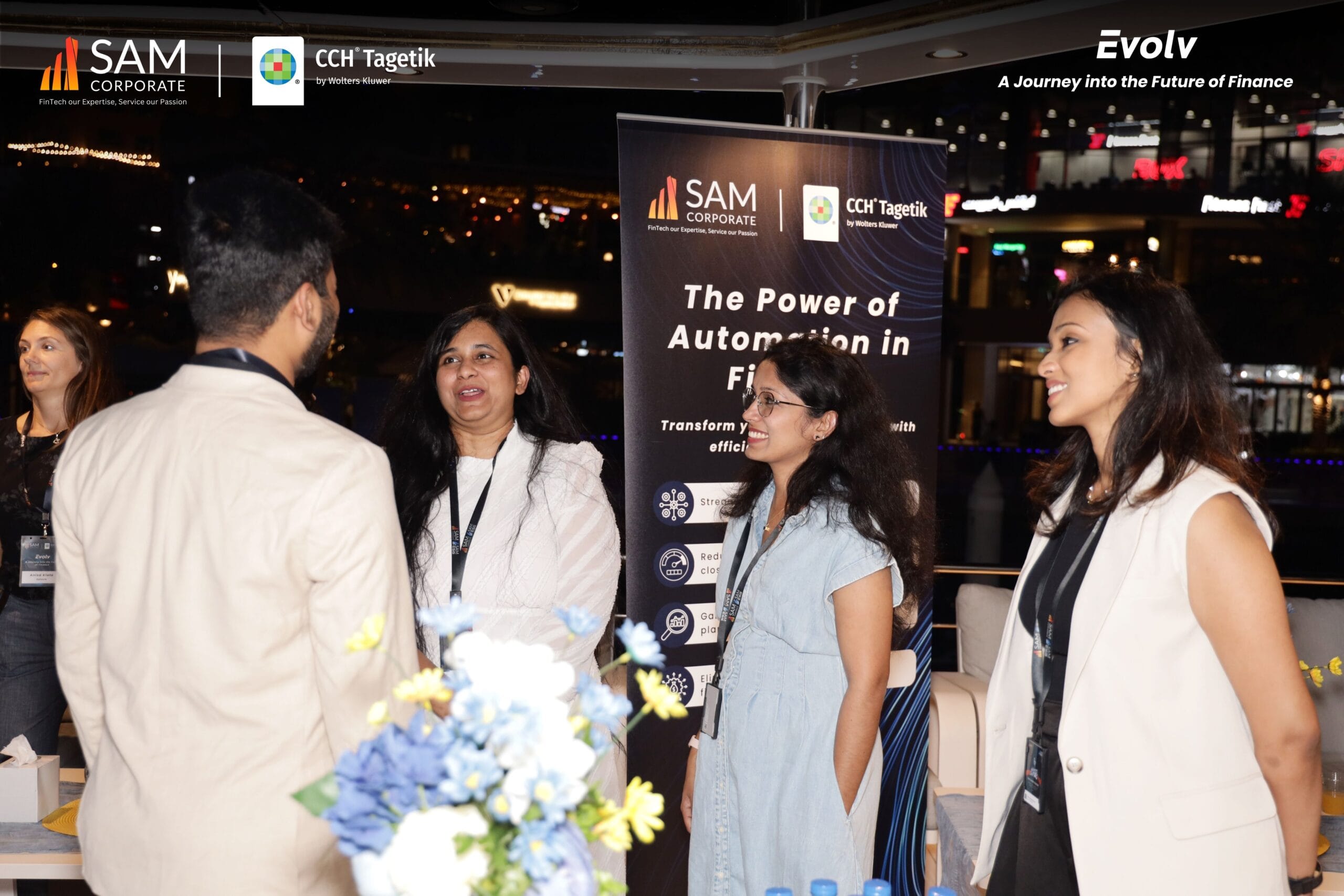 Attendees discussing 'The Power of Automation in Finance' next to a banner at the Evolv event by SAM Corporate and CCH Tagetik.