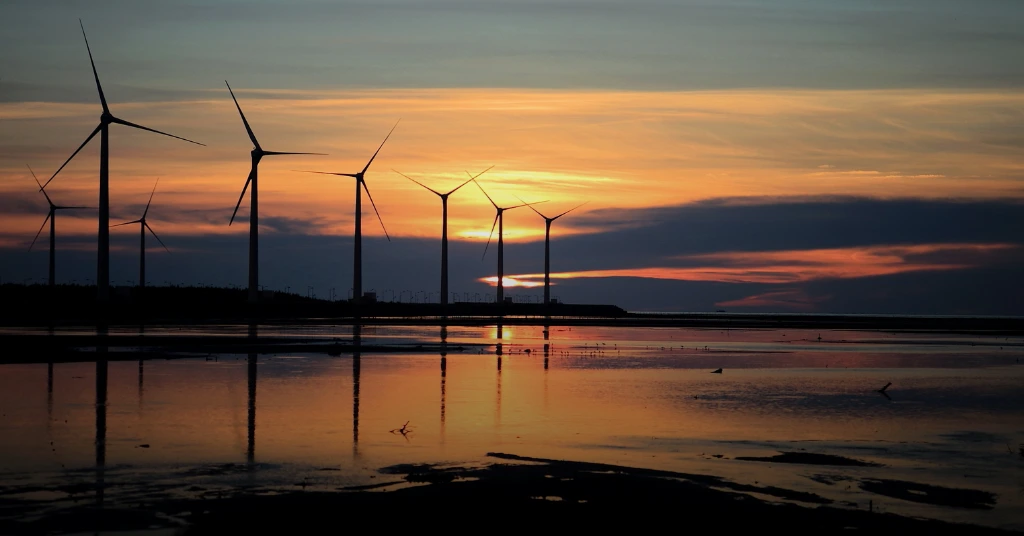 A row of wind turbines silhouetted against a vibrant orange and purple sunset sky, with their reflections visible in the calm water in the foreground.