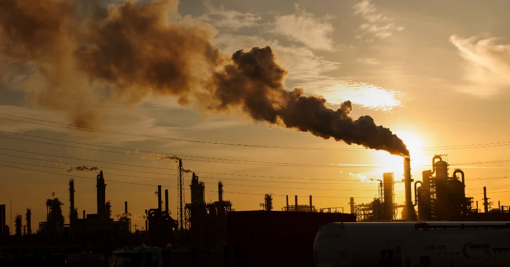 Silhouette of an industrial refinery and storage tanks with smoke rising against a bright orange sunset.