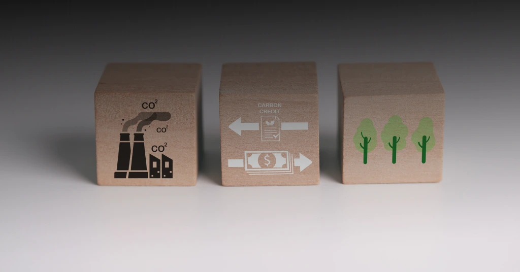 Three wooden blocks showing icons of industrial emissions, a carbon credit transaction with money, and green trees representing carbon sequestration.