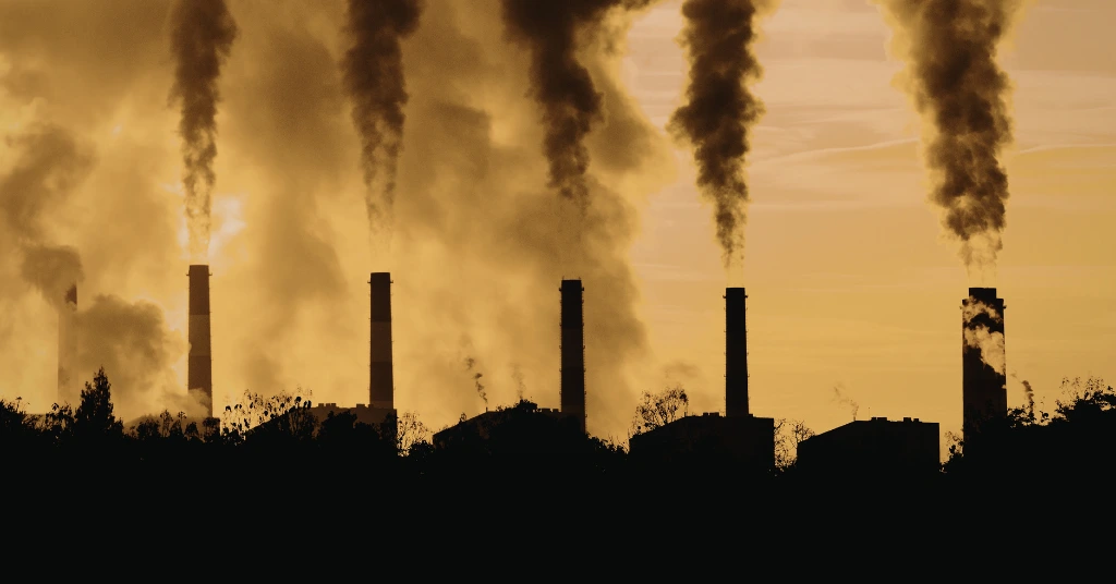 Silhouette of several industrial smokestacks at sunset or sunrise, releasing thick plumes of dark smoke into the hazy atmosphere.