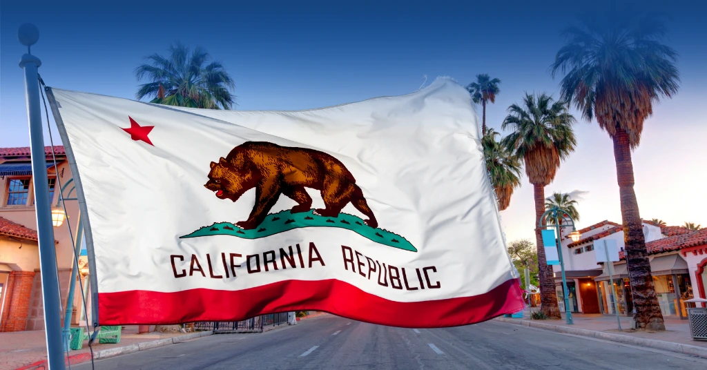 The California state flag waving on a pole in an outdoor urban setting with palm trees and buildings in the background.