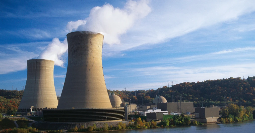 A landscape view of nuclear power plant cooling towers and reactor domes situated along a river with autumn trees in the background.