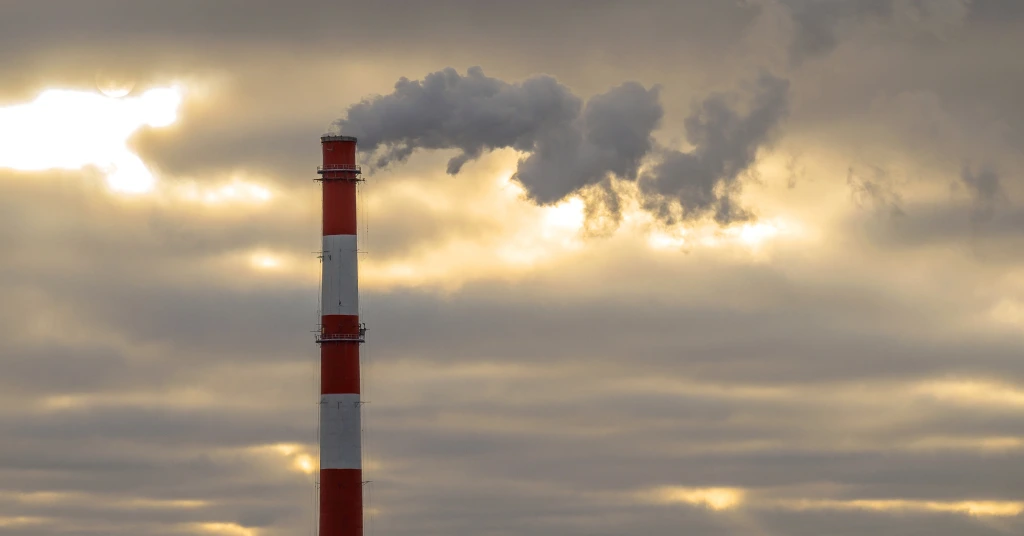 A red and white industrial chimney stack releasing dark smoke into a cloudy sunset sky.